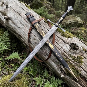 May include: A double-edged steel sword with a patterned blade and a black hilt featuring gold accents. The sword rests on a matching black scabbard decorated with gold leaf-like engravings and a brown leather strap, resting on a mossy log.