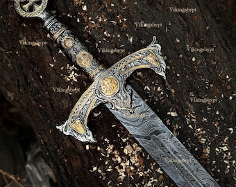 May include: An ornate sword with a silver and gold hilt and a patterned blade. The hilt features intricate designs and a cross-shaped pommel. The blade has a distinctive wavy pattern. The sword rests on a wooden surface.