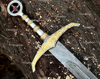 May include: A detailed image of a sword with a silver blade and a gold-colored hilt. The hilt features intricate designs and a red gem. The sword is resting on a piece of wood, showcasing its craftsmanship and historical aesthetic.