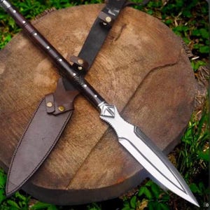 May include: A handcrafted spear with a dark wooden shaft and a silver blade. The spear is paired with a brown leather sheath and a black leather strap. The spear is resting on a wooden surface.