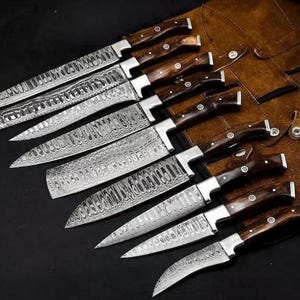 Op de afbeelding: Een assortiment Damascus stalen messen met donkerhouten handvatten. De messen hebben een uniek patroon. De messen zijn gerangschikt op een donkere ondergrond, met een bruine leren schede op de achtergrond.