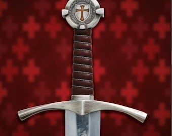 May include: A medieval sword with a silver cross-shaped hilt and a brown leather-wrapped handle. The blade is silver and features an inscription. The sword's pommel has a shield design with a cross. The background is a red pattern.