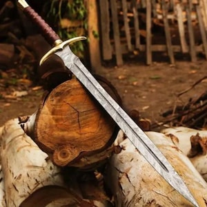Könnte beinhalten: Ein langes Schwert mit gemusterter Klinge und goldenem Parierstück. Der Griff ist mit einem dunkelbraunen Material umwickelt. Das Schwert liegt auf einem Stapel geschnittener Baumstämme, mit einem verschwommenen Hintergrund aus Holzstrukturen.