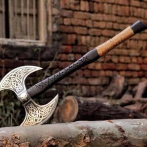 Op de afbeelding: Een zilveren en zwarte dubbelbladige bijl met ingewikkeld Keltisch knoopwerk op de bladen. De handgreep is omwikkeld met bruin leer en zwarte accenten. De bijl rust op een houten stam.