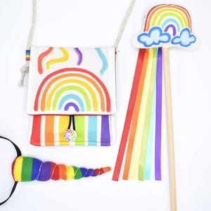 May include: A white crossbody bag with a rainbow print, a matching wand featuring a plush rainbow and cloud topper with long colorful ribbons, and a spiral rainbow horn accessory with a black elastic band, all featuring vibrant multi-colored designs.