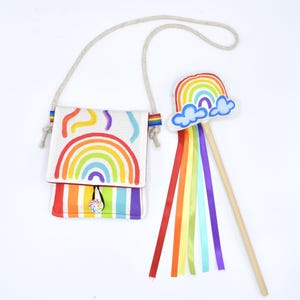 May include: A white crossbody bag featuring a colorful rainbow graphic, a striped flap, and a spiral button closure, paired with a matching wand topped with a rainbow and cloud cutout, featuring long, flowing rainbow-colored satin ribbons.