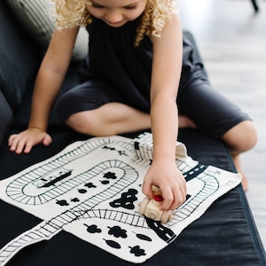 May include: A white fabric play mat featuring black printed illustrations of train tracks, trees, and a tunnel, paired with a small wooden toy train engine with a red button on top, designed for interactive play on a flat surface.