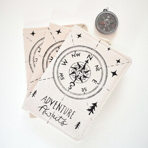 May include: Three off-white fabric passport covers featuring a black printed compass design, small stars, pine trees, and the text "Adventure Awaits." A circular metal compass with a black face and white markings is positioned above the covers.