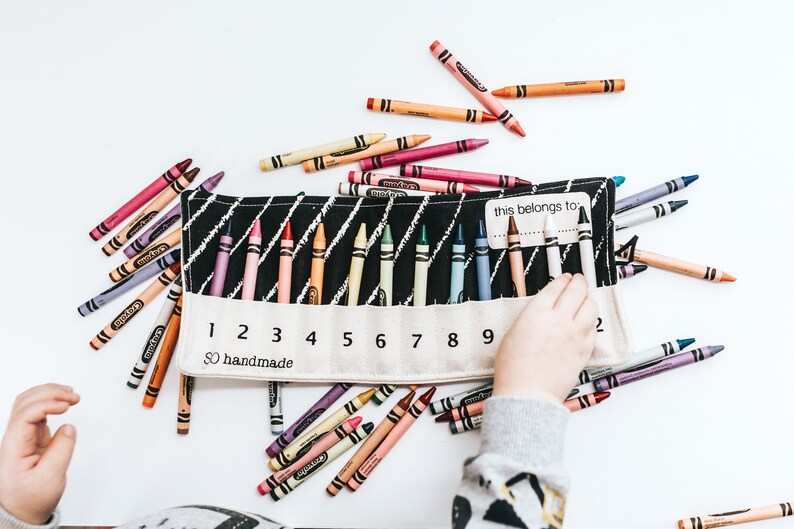 May include: A white canvas crayon roll with black stripes and numbers 1 through 9. The roll is filled with crayons and has a label that says "this belongs to".