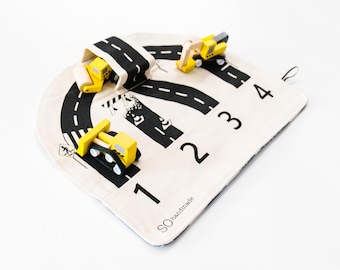 May include: A cream-colored fabric play mat featuring a black printed road design, traffic signs, and the numbers 1, 2, 3, and 4. Three small yellow and black wooden construction vehicles, including a bulldozer and excavator, are placed on the mat.