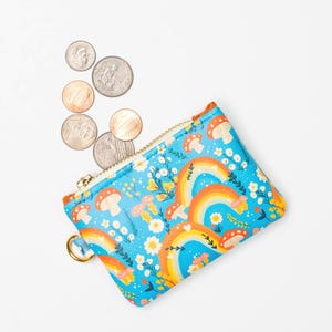 May include: A rectangular zippered coin pouch featuring a bright blue background with a repeating pattern of orange rainbows, white flowers, and red mushrooms. A gold-toned metal zipper and a small circular metal ring are attached to the side.