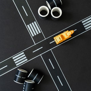May include: A yellow toy truck on a black surface with white tape forming a road intersection. There are several black and white striped paper tubes on the surface.