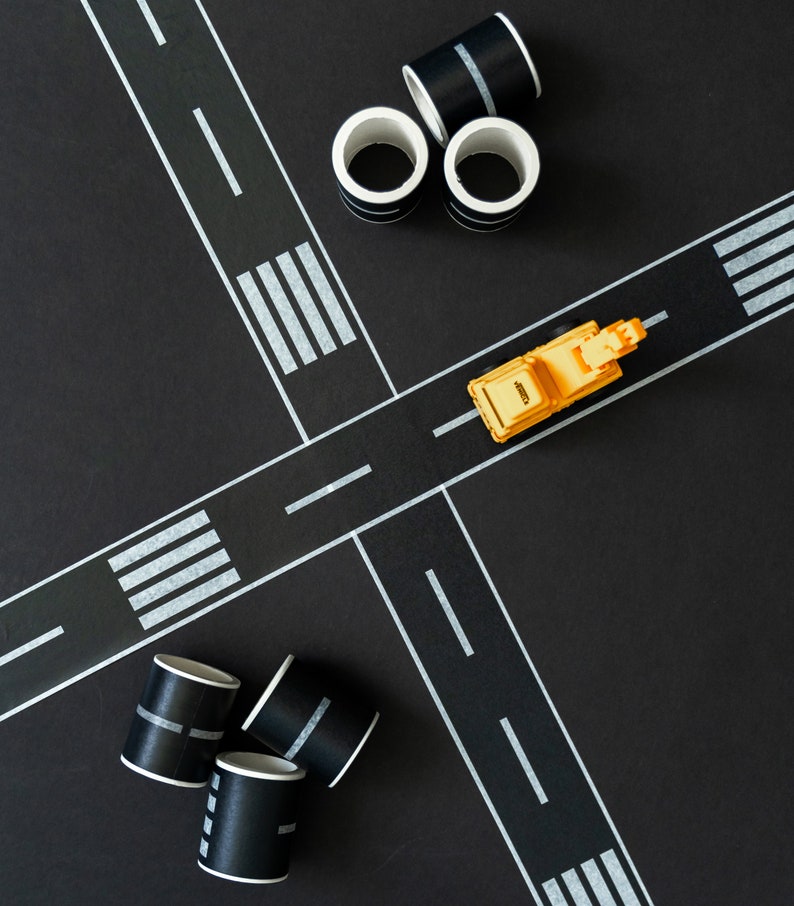 May include: A yellow toy truck on a black surface with white tape forming a road intersection. There are several black and white striped paper tubes on the surface.