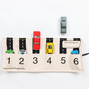 May include: A beige fabric toy car organizer with six numbered slots, each featuring a black road graphic with white dashed lines. The organizer includes a label area with the text 'this belongs to:' and a black drawstring. Five toy cars are placed in the slots, with one grey car positioned above the organizer.