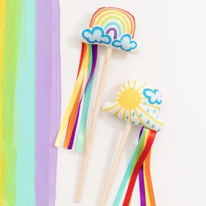 May include: Two fabric wands on wooden sticks. One wand features a plush rainbow with blue clouds and long, multi-colored satin ribbons. The other wand features a plush sun and cloud design with blue raindrops and long, multi-colored satin ribbons.