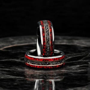 Könnte beinhalten: Zwei Ringe mit einem schwarzen Band, roten Edelstein-Inlays und einem Mittelteil aus dunkelgrauem Stein. Ein dünnes Goldband akzentuiert die roten und grauen Abschnitte. Die Ringe werden auf einer dunklen, reflektierenden Oberfläche präsentiert.
