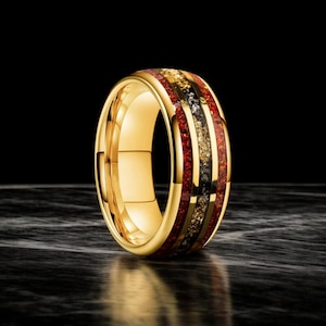 Puede incluir: Un anillo dorado con una banda de incrustaciones rojas, negras y doradas. El anillo tiene un acabado pulido y está sobre un fondo oscuro. El anillo es una pieza de joyería.