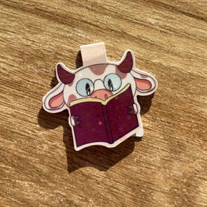 Può includere: Un segnalibro a forma di mucca, con corpo rosa chiaro e bianco, corna marroni e occhiali, che legge un libro bordeaux con una stella. Il segnalibro ha una piccola linguetta per un facile posizionamento in un libro. Accessorio divertente.