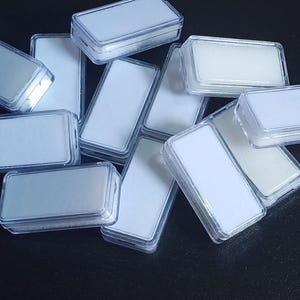 Può includere: Una collezione di piccole scatole rettangolari di plastica trasparente con inserti bianchi. Le scatole sono impilate e sparse, con alcuni coperchi aperti, rivelando l'interno bianco. L'immagine è ripresa da un'angolazione leggermente elevata, su sfondo scuro.