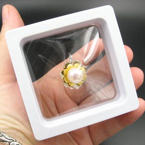 May include: A flower-shaped pendant with a pearl centre, held in a white display case. The pendant has gold-coloured petals and silver-coloured accents. The pearl is a creamy white colour. The display case is square with a clear plastic window.
