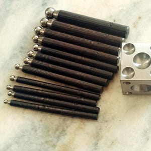 May include: A collection of dark brown tools with silver ball tips and a silver metal cube with rounded indentations. The tools are arranged in a diagonal pattern, suggesting a set for a specific craft or repair.