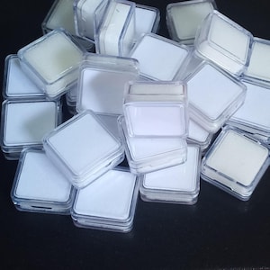 Pode incluir: Uma coleção de pequenas caixas quadradas de plástico transparente, cada uma com um interior branco e uma inserção de espuma. As caixas estão empilhadas e espalhadas sobre uma superfície escura, sugerindo um tema de armazenamento ou exibição.