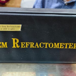 May include: A black box with the words "Gem Refractometer" in gold lettering. A smaller label above reads "RHG-151 Gem Refractometer Build in LED Light". The box is sitting on a light-colored surface.