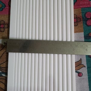 May include: A close-up of a white, ridged material with a metal ruler across it. The ruler displays measurements in centimetres, showing the material's width. A patterned fabric forms the background.