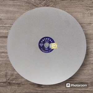 May include: A circular light gray diamond grinding disc with a textured surface. The center features a dark blue circular label with the text Crystal Gold, Diamond Disc, and the number 600, along with a small gold holographic sticker.