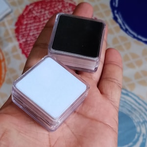 Pode incluir: Duas caixas de joias quadradas de plástico transparente, uma com uma inserção preta e outra com uma inserção branca, são seguradas em uma mão. As caixas têm tampas transparentes. O fundo é um tecido colorido estampado.