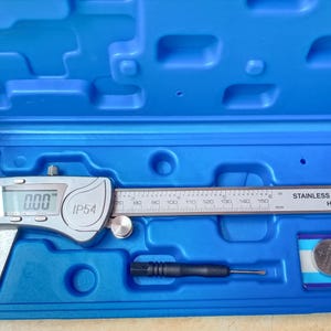 Può includere: Un set di calibri digitali in una custodia di plastica blu. Il calibro argentato ha un display digitale che mostra "0.00". La custodia contiene anche un piccolo cacciavite e una batteria al litio CR2032. La scala di misurazione è in millimetri.