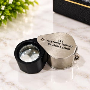 May include: A Bausch & Lomb 14X Hastings Triplet jeweller's loupe featuring a black circular lens housing attached to a brushed silver metal swing-away cover. The silver cover is engraved with the text 14X HASTINGS TRIPLET BAUSCH & LOMB.
