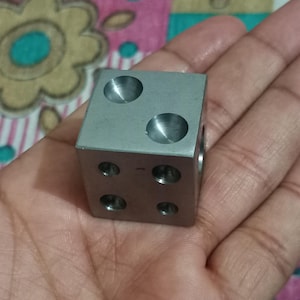 Puede incluir: Un pequeño cubo de metal plateado con seis agujeros circulares perforados en sus superficies. El cubo se sostiene en una mano y los agujeros están espaciados uniformemente. El cubo parece ser una pieza mecanizada de precisión.