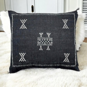 May include: A square, dark gray decorative pillow with white embroidered geometric patterns. The pillow has a fringed edge and is placed on a white, fluffy surface. The embroidered design includes X-shaped and rectangular motifs.