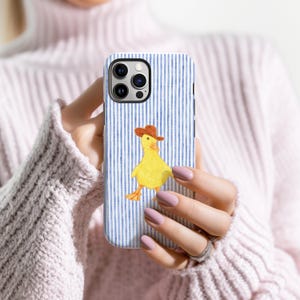 May include: A phone case with a blue and white striped background featuring a yellow duck wearing a brown cowboy hat. The phone case is held in a person's hands.