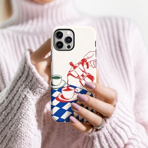 May include: A white phone case with a red and blue illustration of a coffee pot pouring coffee into a cup. The design includes a blue and white checkered pattern and a green cup. The case is held in a person's hands.