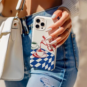May include: A white phone case featuring a coffee-themed illustration. The design includes a red and white coffee pot pouring coffee into a cup on a blue and white checkered tablecloth. A white purse and blue jeans are also visible.