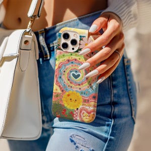 Puede incluir: Una funda de teléfono colorida con un diseño caprichoso con un sol, flores y un corazón. La funda tiene un patrón de patchwork con la palabra "CHANGE" visible. La funda la sostiene una persona que lleva vaqueros azules y un bolso blanco.