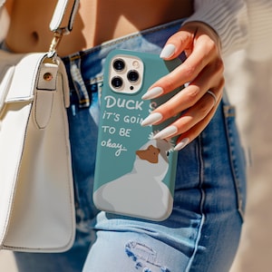 May include: A teal phone case with a cartoon duck design and the words "Duck's it's goin' to be okay." The case shows a white duck with a brown beak. The phone case is held by a person wearing blue denim jeans.