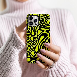 K&ouml;nnte beinhalten: Eine Handyh&uuml;lle mit einem neon-gelben und schwarzen Muster. Das Case hat ein sich wiederholendes abstraktes Design aus schwarzen organischen Formen auf einem leuchtend gelben Hintergrund. Die Handyh&uuml;lle wird von einer Person gehalten.