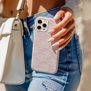 May include: A light pink phone case with a delicate lace pattern. The case has a camera cutout and is held by a person wearing blue jeans and a white top. A white purse with a gold strap is also visible.