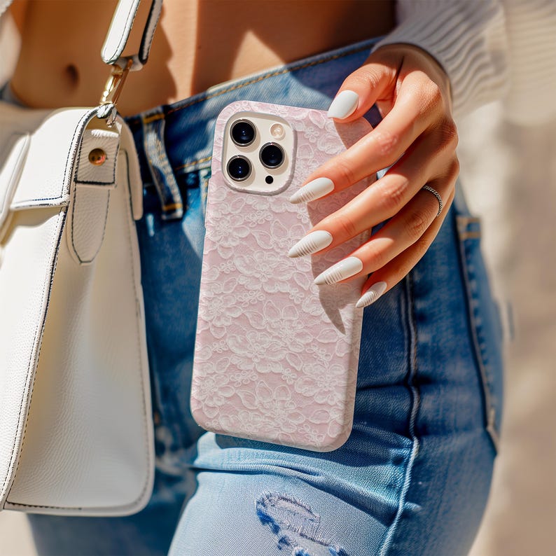 May include: A light pink phone case with a delicate lace pattern. The case has a camera cutout and is held by a person wearing blue jeans and a white top. A white purse with a gold strap is also visible.