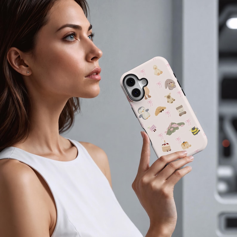 May include: A light pink phone case featuring a repeating pattern of cute animal illustrations, including a bunny, penguin, and bear. The case has a black border and is held by a person.