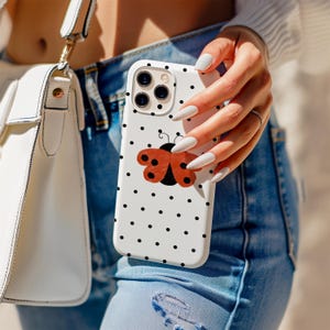 May include: White phone case with black polka dots and a red and black ladybug design. The phone case is held in front of a person wearing blue jeans and a white purse.