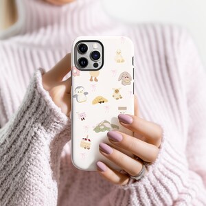 May include: A white phone case featuring a repeating pattern of cute cartoon animals and pink bows. The case has a camera cutout and is held in a person's hands. The animals include a penguin, duck, bunny, and bear.