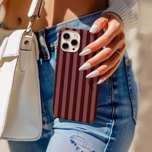 Pode incluir: Uma capa de telefone listrada em bordô e marrom. A capa é segurada em frente a uma pessoa usando jeans azuis e uma bolsa branca. A capa do telefone tem uma câmera de lente tripla.