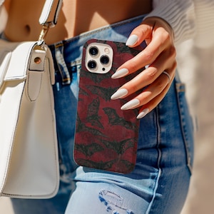 May include: A burgundy phone case featuring a pattern of black bats. The case is held by a person wearing blue jeans. A white purse with a gold strap is visible in the background.