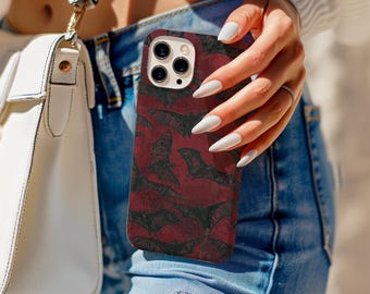 May include: A burgundy phone case featuring a pattern of black bats. The case is held by a person wearing blue jeans. A white purse with a gold strap is visible in the background.