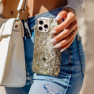 Puede incluir: Funda de teléfono decorativa con un diseño de mosaico en tonos plateados y dorados. La funda presenta un acento en forma de estrella y un recorte para la cámara. La persona lleva vaqueros azules y un suéter blanco.