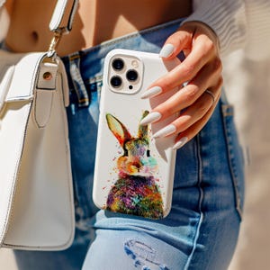 May include: White phone case with a vibrant watercolour rabbit design. The rabbit is depicted with a rainbow-coloured body and ears. The phone case is held by a person wearing blue jeans and a white sweater.